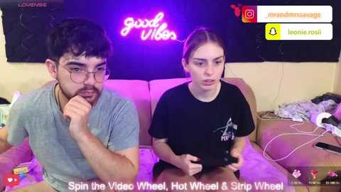 Video of a young man and woman playing a video game on a couch, with neon \"good vibes\" sign and gaming peripherals in the background.