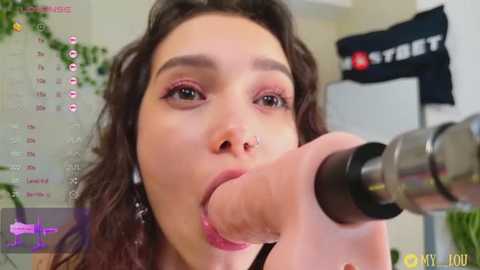 Media: Video of a young woman with wavy brown hair, light skin, and pink lipstick, performing oral sex on a large, realistic sex toy. The background features a calendar, plants, and a \"STAR\" banner.