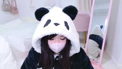 Media: Video of a young Asian woman with long dark hair wearing a black and white panda onesie and a pink face mask, sitting on a white bed in a bright, minimalistic room.