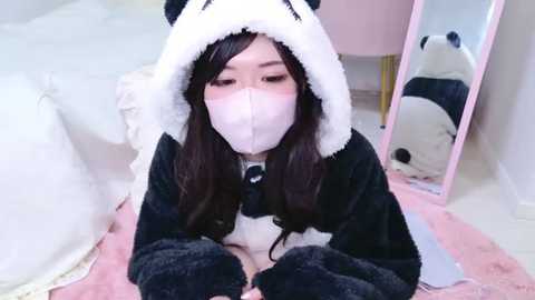 Media: Video of an Asian woman with long black hair, wearing a panda onesie, face mask, and panda hood, sitting on a pink rug in a soft, pastel-colored room.