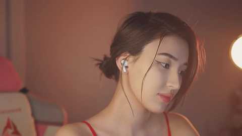 Media: Video of a young Asian woman with dark hair in a bun, wearing red spaghetti straps, listening to white earbuds, in a dimly lit, warmly-colored room with a round light source.