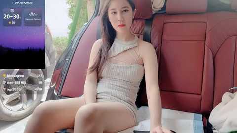 Media: Video of a young East Asian woman with long black hair, fair skin, and red lipstick, wearing a sheer, beige mini dress, sitting in a red car.