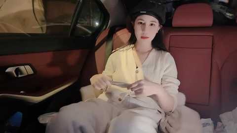 Media: A video of a young woman with pale skin and straight black hair, wearing a beige cardigan and a black beanie, seated in the back of a vintage car with red leather seats, adjusting a small white object in her lap.