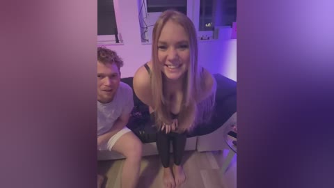 Media: Video of a young woman with long blonde hair, leaning forward with a smile, and a man with curly hair and a bandage on his arm, sitting on a wooden floor in a dimly lit room with purple lighting.