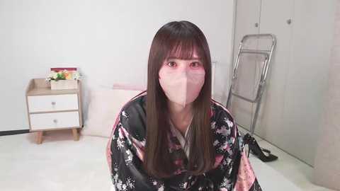 Media: Video of a young woman with long, straight brown hair, wearing a floral kimono, white mask, and sitting on a bed in a minimalist room with white walls, a wooden nightstand, and a metal ladder.