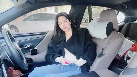 Media: Video of an Asian woman with long black hair, wearing a black jacket and jeans, sitting in a beige leather car seat. She appears relaxed and is holding a small white bag.