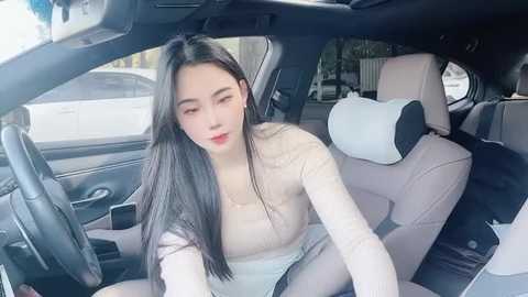 Media: Video of a young East Asian woman with long black hair, fair skin, and red lipstick, wearing a beige top, sitting in a car with beige seats.