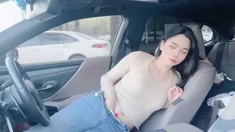 Video of a young woman, possibly in her 20s, with long black hair, wearing a beige top and blue jeans, slouched in the front passenger seat of a car, eyes closed, looking tired.