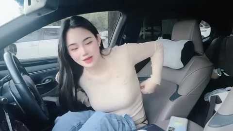 Media: Video of an Asian woman with long black hair, wearing a beige top, blue jeans, and white sneakers, sitting in the back seat of a car, looking relaxed.