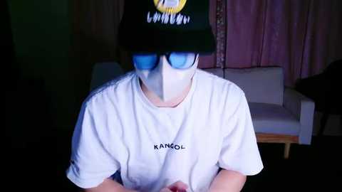 Media: Video of a person in a white Kangol t-shirt and black Kangol cap, wearing blue-tinted sunglasses and a white mask, indoors with pink curtains and a white couch in the background.