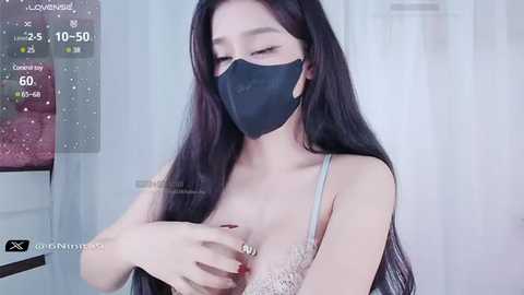 Media: Video of an East Asian woman with long black hair, wearing a black face mask and beige lace bra, indoors with white curtains and a digital clock showing 25.5\u00b0C.