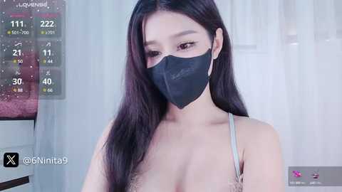 Media: A video of a young Asian woman with long black hair and fair skin, wearing a black face mask, a light gray bra, and a necklace. Background shows a calendar and a window.