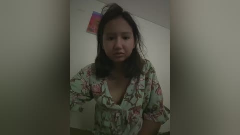 Media: A video of an Asian woman with shoulder-length dark hair, wearing a floral-patterned robe, standing in a dimly lit hallway. The background features plain beige walls with a colorful sticker near the top left corner.