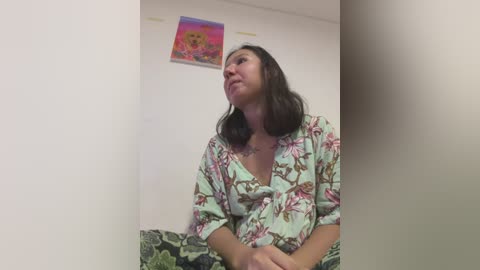 Media: Video of a light-skinned woman with shoulder-length dark hair, wearing a floral-patterned robe, seated on a green-patterned couch, looking upward, against a plain white wall with a colorful abstract painting above.