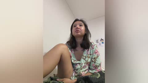 Media: Video of a young woman with medium skin tone, dark hair, and a floral blouse, sitting in a corner of a plain white room.