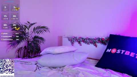 A video of a minimalist bedroom with a white bed, a green potted plant, a floral garland, and a large \"MOTSEBE\" TV screen. The room has a purple ambient light.