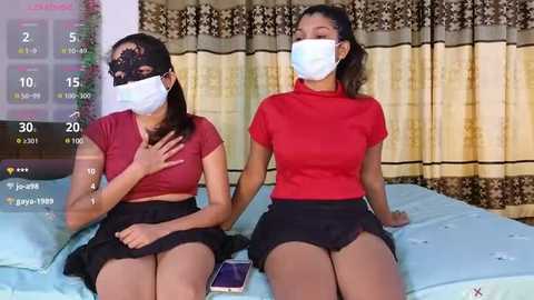 Media: Video of two Asian women in face masks, red tops, and black skirts, sitting on a bed with blue sheets, in a room with yellow curtains.