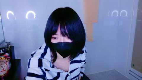 Media: Video of an East Asian woman with straight black hair, wearing a black face mask, white striped shirt, and beige top, sitting indoors near a TV and snacks.