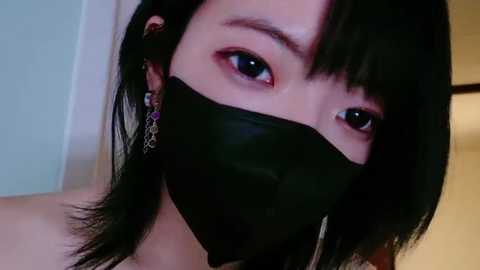 Media: Close-up video of an Asian woman with short black hair, wearing a black face mask, showing her large, almond-shaped eyes and delicate facial features.