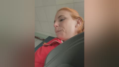 Media: Video of a red-haired woman with fair skin wearing a red jacket, seated in a dimly lit, tiled room with a ceiling grid.