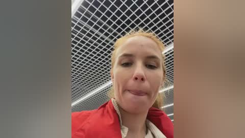 Media: Video of a blonde woman with fair skin, wearing a red jacket, in a modern, industrial space with a grid-patterned ceiling. She has a neutral expression.