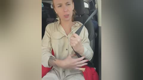 Media: Video of a blonde woman in beige overalls, holding steering wheel, seated in a car with red seatbelt.