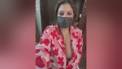 Media: A video of a woman with light brown skin, wearing a red floral-patterned robe with a deep V-neckline, a gray face mask, and a gold necklace. She has medium-length brown hair and sits in a dimly lit room with brown curtains.