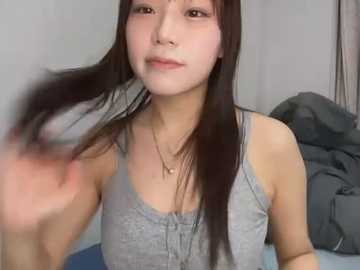 Media: A video of an East Asian woman with long black hair, wearing a grey tank top, playing with her hair in a bedroom with grey walls and a blue bed.