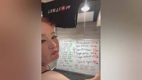 Media: Video of a fair-skinned woman with red hair, wearing black clothing, reading a whiteboard with detailed notes in green and red markers. The background shows a dark wall and a \"WELCOME\" sign.