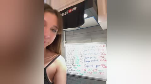 Media: Video of a young woman with long brown hair, wearing a black spaghetti-strap top, in a dimly lit room with a whiteboard covered in red and black handwritten notes.