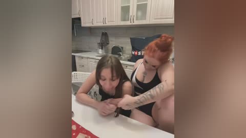 Media: Video of a red-haired woman with tattoos, wearing a black bra, pulling down the black tank top of a brunette woman on a kitchen counter.