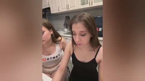 Media: Video of two young women in a kitchen. One wears a white tank top, the other a black tank top. Both have medium build, light skin, and straight brown hair. Background features white cabinets, a toaster, and a coffee maker.