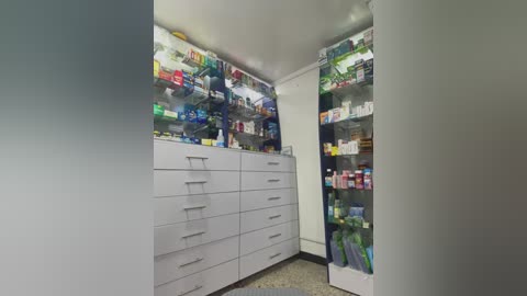 Media: Video of a well-lit, organized pharmacy with white cabinets and shelves stocked with various medications and supplies. The background is a plain white wall, enhancing the clean and sterile environment.