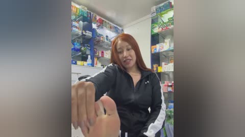 Media: Video of a smiling, red-haired woman in a black jacket, holding out a foot in a pharmacy. Shelves of colorful medicines and products in the background.