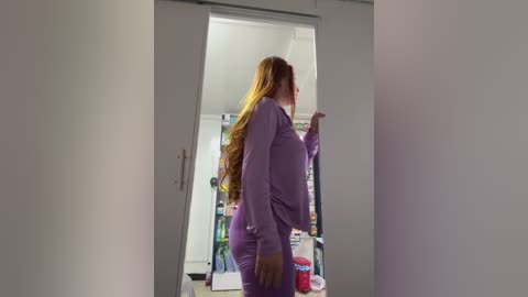 Media: Video of a plus-sized woman with long, wavy red hair, wearing a purple robe and matching pajama pants, standing in front of a mirrored closet door. Behind her, a kitchen with shelves filled with various items is visible.