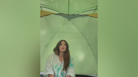 Media: Video of a woman with long brown hair, wearing a white shirt with blue abstract designs, sitting inside a translucent, greenish, tent-like structure with a yellow and black roof.