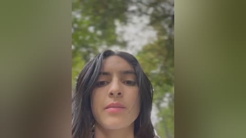 Media: Video of a young woman with long, dark hair, light brown skin, and a neutral expression, standing in a lush, green forest.