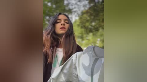 Media: Video of a young woman with long, dark hair, wearing a black jacket over a white shirt, holding a gray and white-patterned cloth against a blurred green background.