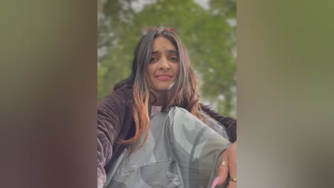 Media: Video of a young woman with long, dark hair, wearing a light blue and white striped shirt, standing outdoors with blurred greenery in the background.