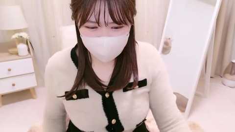 Media: Video of an Asian woman with long brown hair, wearing a white mask, black dress with gold buttons, and a white cardigan. She is indoors, in a softly lit room with white walls, a white dresser, and a mirror.