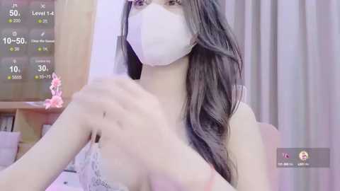 Media: Video of a young East Asian woman with long, wavy brown hair, wearing a white mask, pink lace bra, and a white top, adjusting her bra strap. Background includes a wooden bookshelf, a pink doll, and a green curtain.