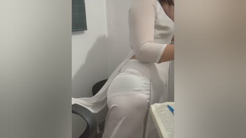 Media: Video of a woman in a white dress, seated, with legs crossed, holding a book, in a bathroom with a gray toilet and white walls.