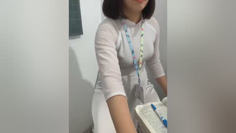 Media: Video of a woman with medium skin tone and shoulder-length dark hair, wearing a white, long-sleeved dress, seated at a small table with an open book and colorful lanyard. Background includes a tiled wall and a window.