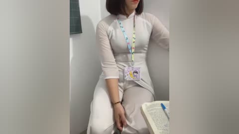 Media: Video of a woman with shoulder-length brown hair, wearing a white lab coat and pants, seated on a bench in a white room. She has a badge around her neck. An open book with notes is on the bench beside her.