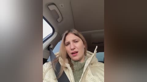 Media: Video of a woman in a car, wearing a green sweater and beige jacket, holding the steering wheel, mouth open, looking upset, inside a modern car with beige interior.