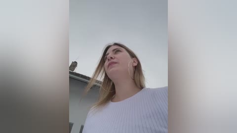 Video of a blonde woman in a white ribbed sweater, looking upward with a contemplative expression, against a blurred, minimalist background.