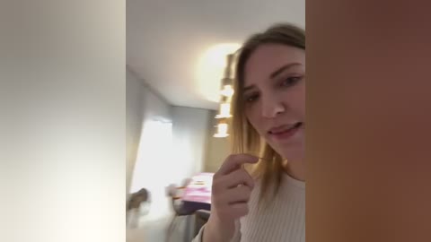 Media: Video of a young, light-skinned woman with long, blonde hair, wearing a beige sweater, standing in a dimly lit, modern kitchen with white walls and pendant lights.