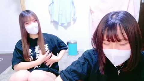 Media: A video shows two young East Asian women in a dimly lit room. Both wear white face masks, black T-shirts, and shorts. The woman on the left, with long straight hair, is seated on the floor, while the one on the right, with shoulder-length hair, is standing.