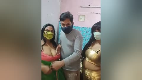 Media: Video of three people, a man and two women, in a small room. The man, in a gray shirt, has a mask and is holding the women's hands. The women wear colorful, revealing outfits.