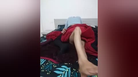 Media: Video of a woman lying on a bed with a black and green floral duvet, wearing a light blue shirt and red skirt, legs crossed.